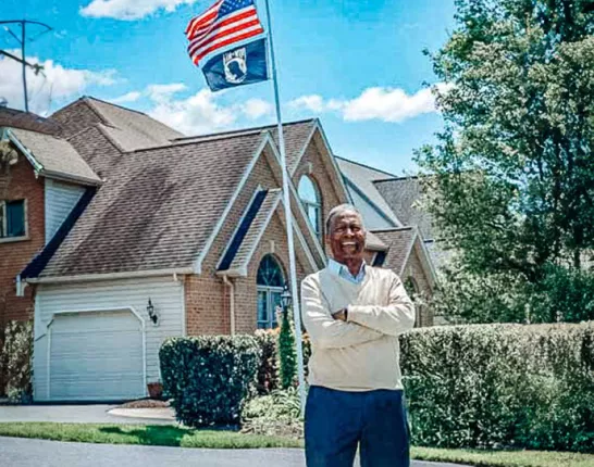 Veteran Jim Williams outside of a house with an American flag and a POW flag
