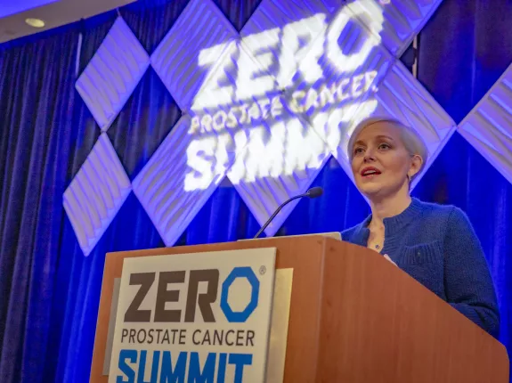 Blonde woman with very short hair standing behind a podium that has a sign saying "ZERO Prostate Cancer Summit"